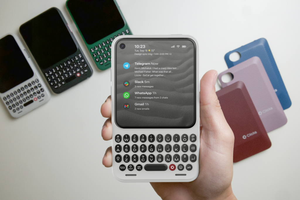 A hand holds a light-colored Clicks Communicator with a physical keyboard and message notifications, beside other phones and colored back covers.