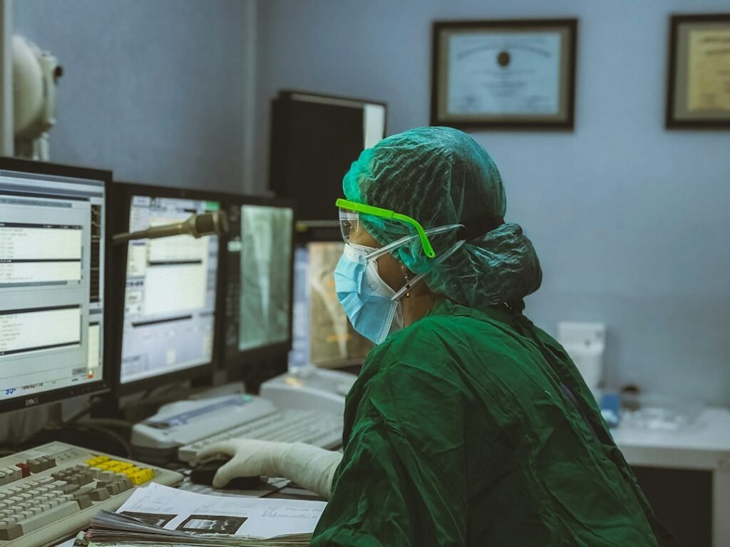 Masked clinician reviewing medical images and patient data on multiple monitors in a hospital control room. Photo by Irwan via Unsplash.