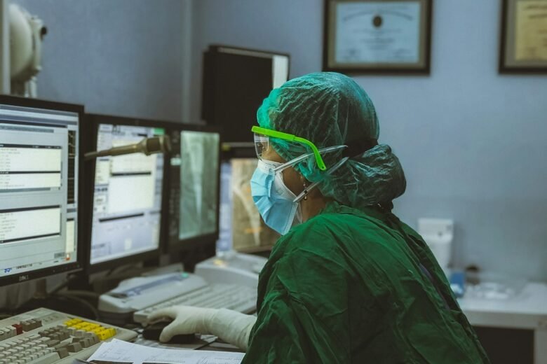 Masked clinician reviewing medical images and patient data on multiple monitors in a hospital control room. Photo by Irwan via Unsplash.