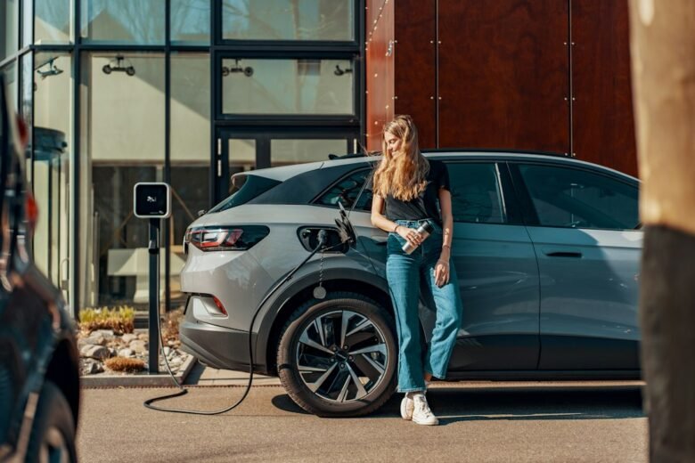 A driver holding a mobile device beside an electric vehicle at a charging station, illustrating phone-based car access. Photo by JUICE on Unsplash.