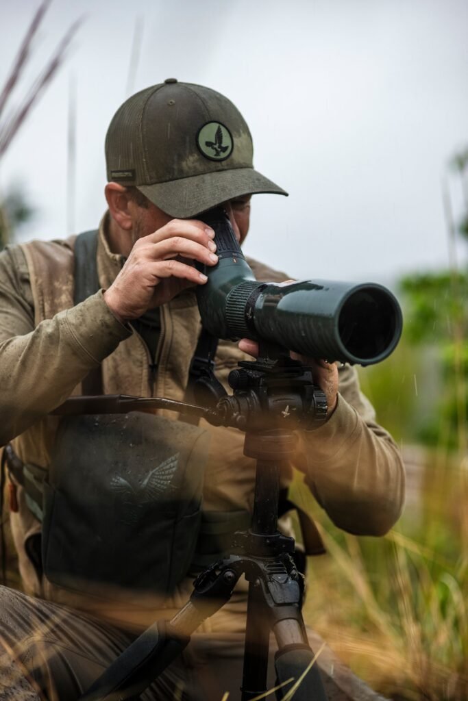 A person uses a green Swarovski AT Endura spotting scope mounted on a tripod in rainy grassland