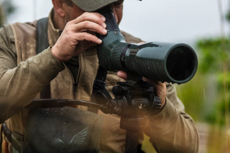 A person uses a green Swarovski AT Endura spotting scope mounted on a tripod in rainy grassland