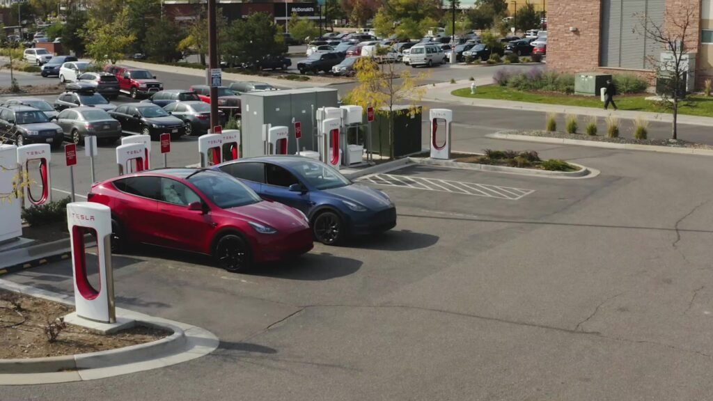 Red and blue Tesla vehicles parked beside white-and-red Supercharger stalls