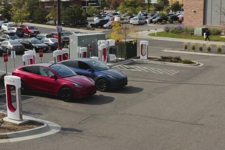 Red and blue Tesla vehicles parked beside white-and-red Supercharger stalls